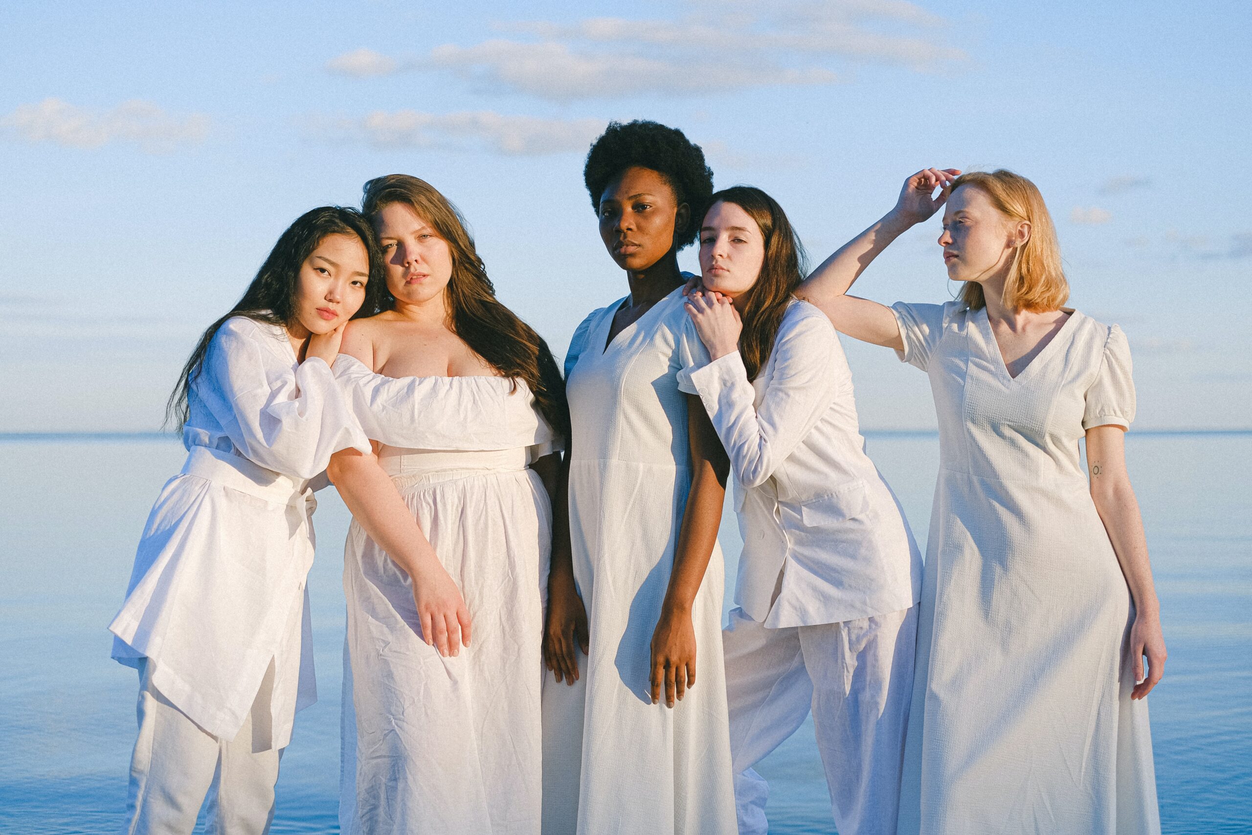 5 women of different color and size stand together dressed in white beside a still lake.